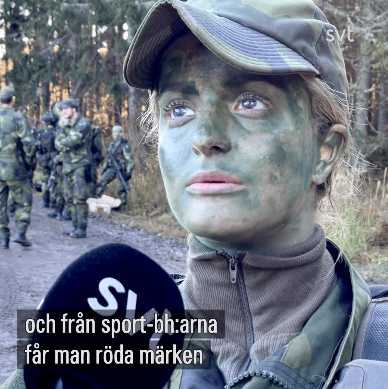 Still from an SVT news video: a conscript in camouflage face paint and a field cap being interviewed with an SVT microphone, soldiers on a forest road behind her