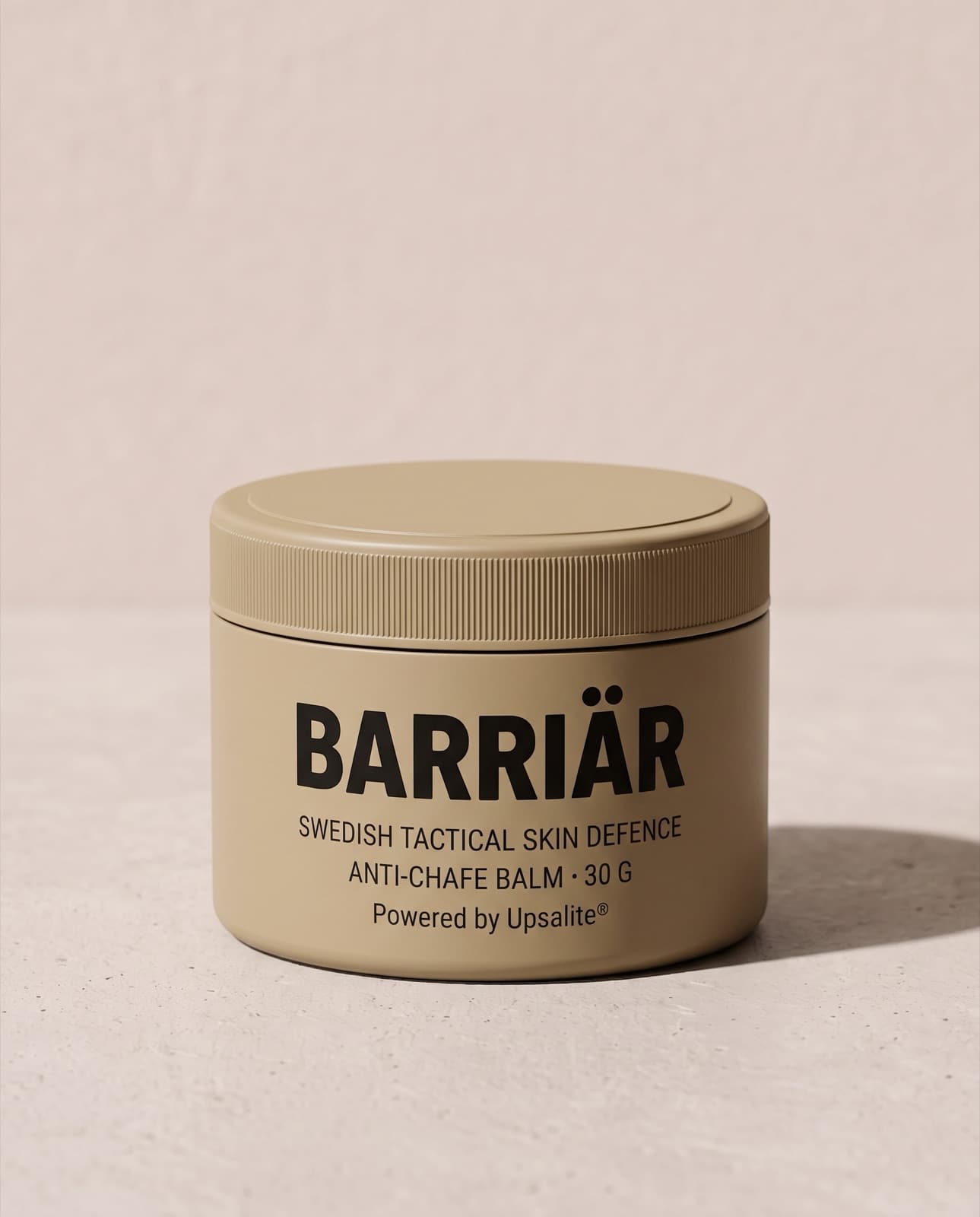 Barriär Anti-Chafe Cream, a wide, low 60 ml sand-beige tin with a knurled screw lid, printed with the Barriär wordmark in Barlow condensed over Swedish Tactical Skin Defence, standing on pale concrete against a plaster wall