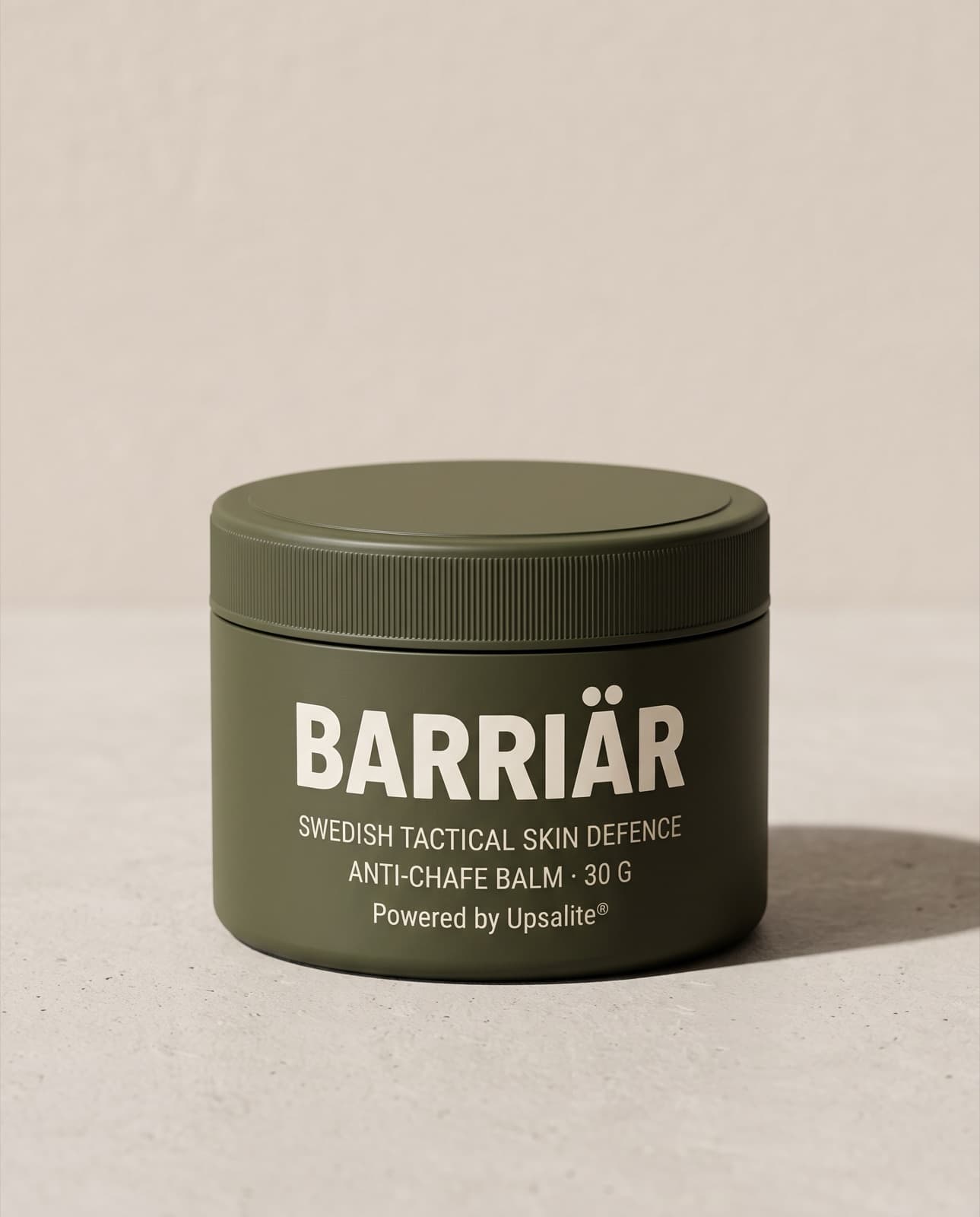 Barriär Anti-Chafe Cream, a wide, low 60 ml olive-green tin with a knurled screw lid, printed with the Barriär wordmark in Barlow condensed over Swedish Tactical Skin Defence, standing on pale concrete against a plaster wall