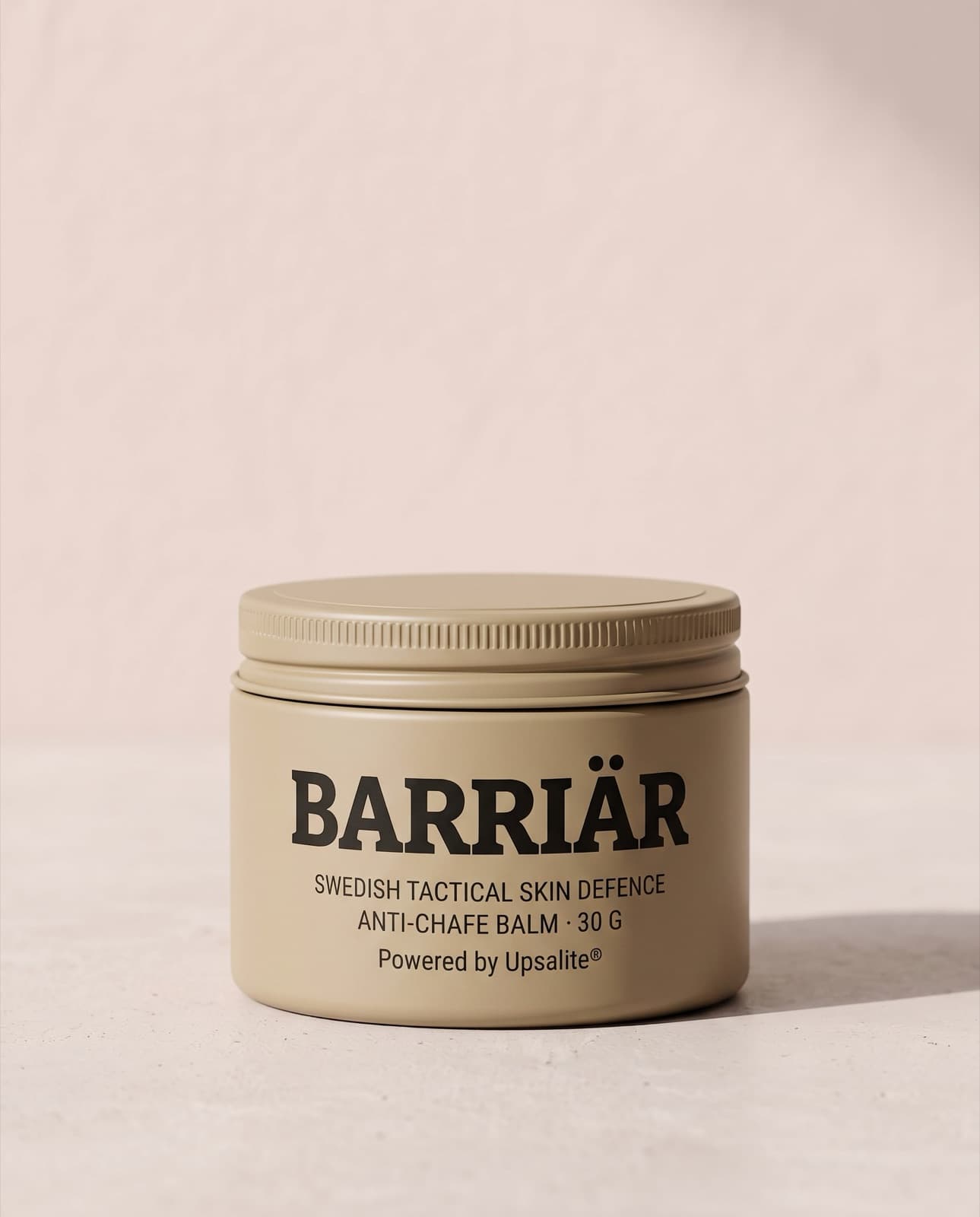 Barriär Anti-Chafe Cream, a wide, low 60 ml sand-beige tin with a knurled screw lid, printed with the Barriär wordmark in Bitter slab-serif over Swedish Tactical Skin Defence, standing on pale concrete against a plaster wall