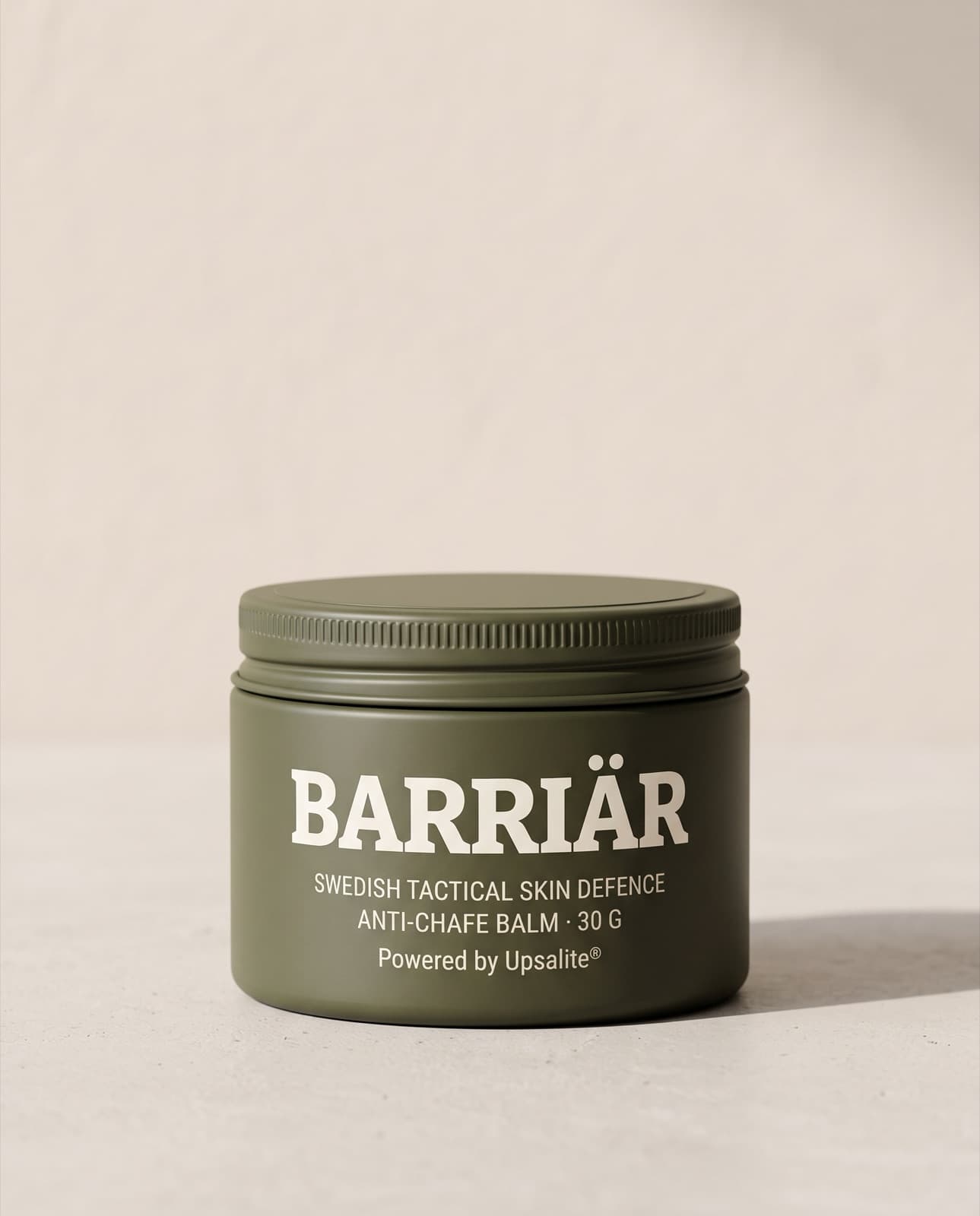 Barriär Anti-Chafe Cream, a wide, low 60 ml olive-green tin with a knurled screw lid, printed with the Barriär wordmark in Bitter slab-serif over Swedish Tactical Skin Defence, standing on pale concrete against a plaster wall