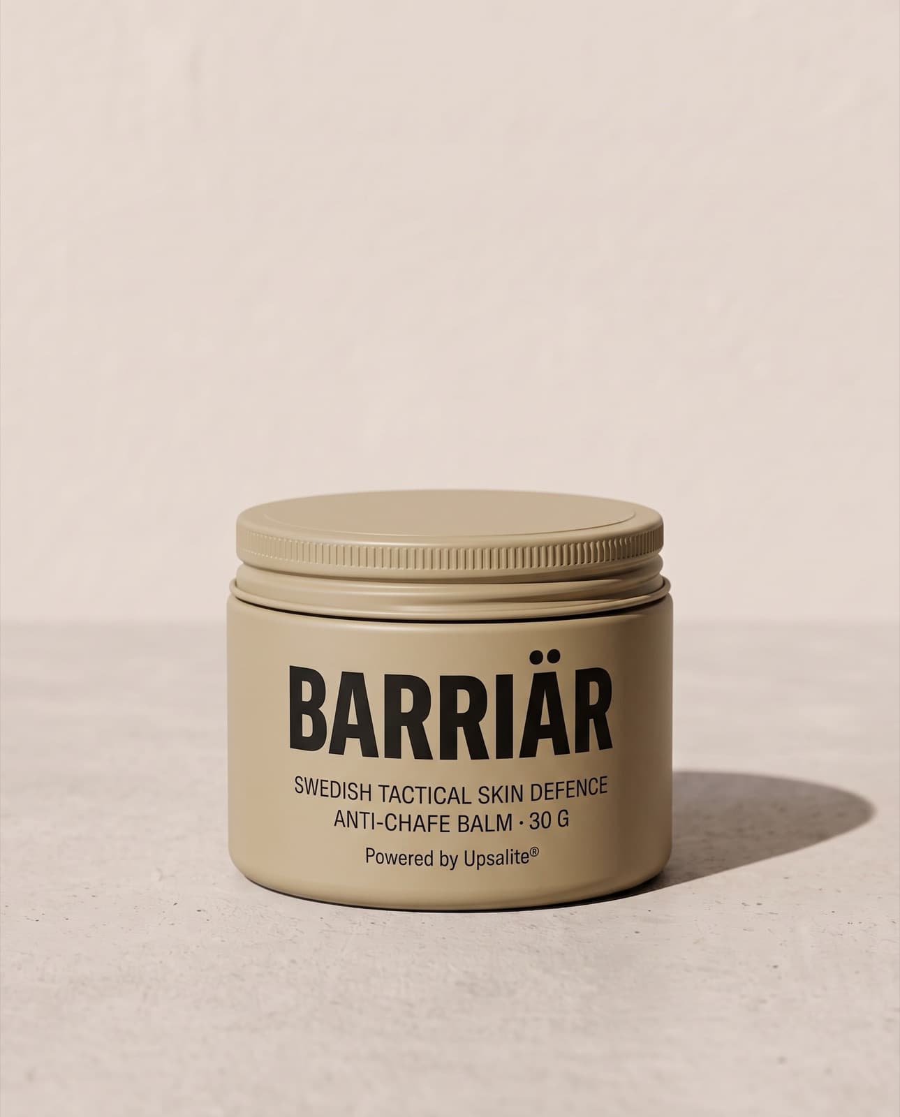 Barriär Anti-Chafe Cream, a wide, low 60 ml sand-beige tin with a knurled screw lid, printed with the Barriär wordmark in Saira condensed over Swedish Tactical Skin Defence, standing on pale concrete against a plaster wall