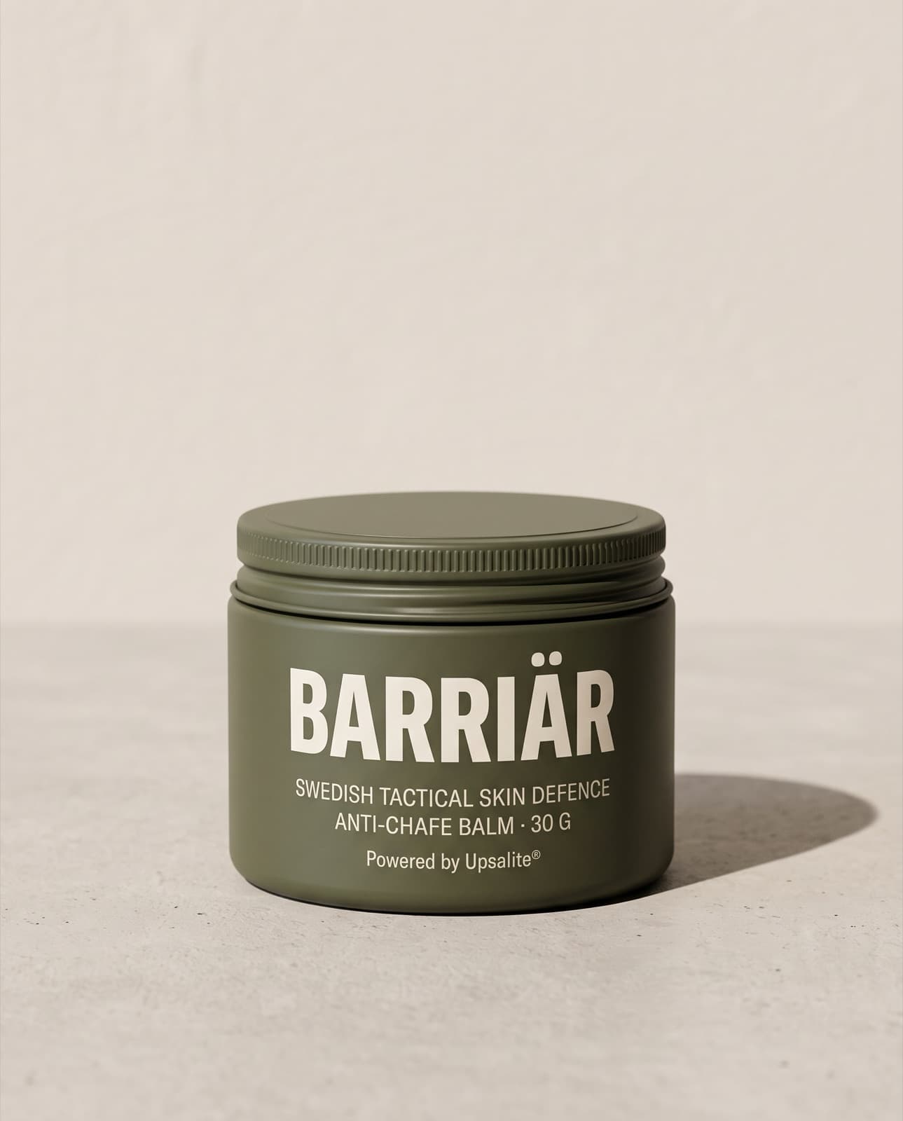 Barriär Anti-Chafe Cream, a wide, low 60 ml olive-green tin with a knurled screw lid, printed with the Barriär wordmark in Saira condensed over Swedish Tactical Skin Defence, standing on pale concrete against a plaster wall