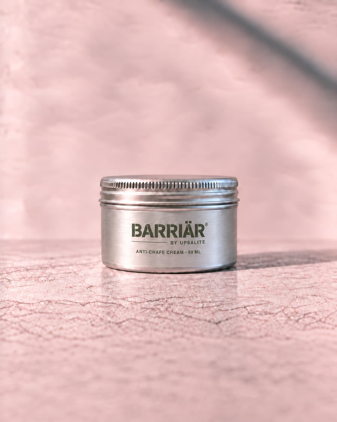 Barriär Anti-Chafe Cream, a wide, low 60 ml brushed aluminium tin with a knurled screw lid, printed in olive straight onto the metal with the Barriär by Upsalite wordmark and Anti-Chafe Cream 60 ml, standing on pale concrete against a plaster wall