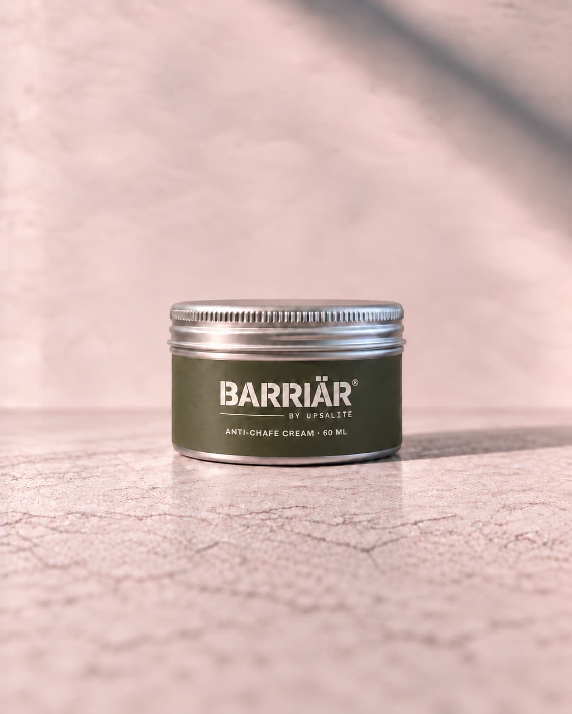 Barriär Anti-Chafe Cream, a wide, low 60 ml brushed aluminium tin with a knurled screw lid and an olive sticker round its side, printed in white with the Barriär by Upsalite wordmark and Anti-Chafe Cream 60 ml, standing on pale concrete against a plaster wall