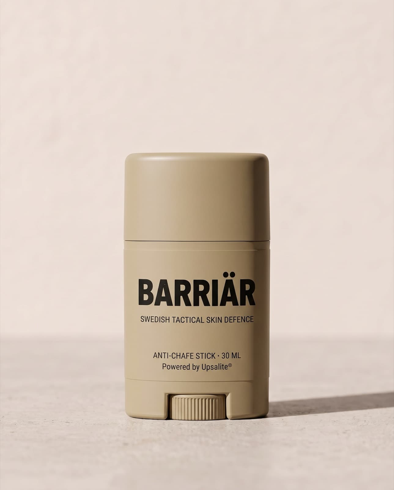 Barriär Anti-Chafe Stick, a short 25 ml sand-beige twist-up stick printed with the Barriär wordmark in Barlow condensed over Swedish Tactical Skin Defence, standing on pale concrete against a plaster wall
