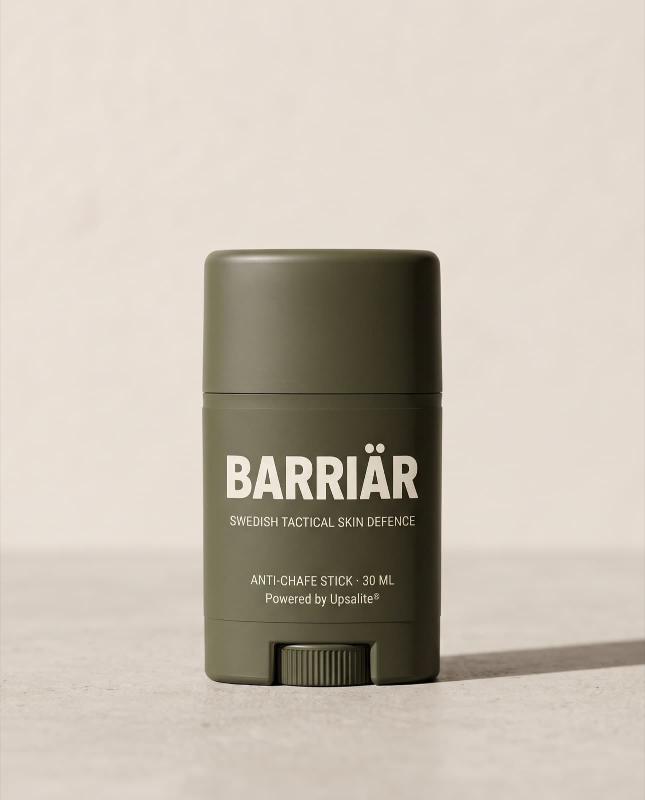 Barriär Anti-Chafe Stick, a short 25 ml olive-green twist-up stick printed with the Barriär wordmark in Barlow condensed over Swedish Tactical Skin Defence, standing on pale concrete against a plaster wall
