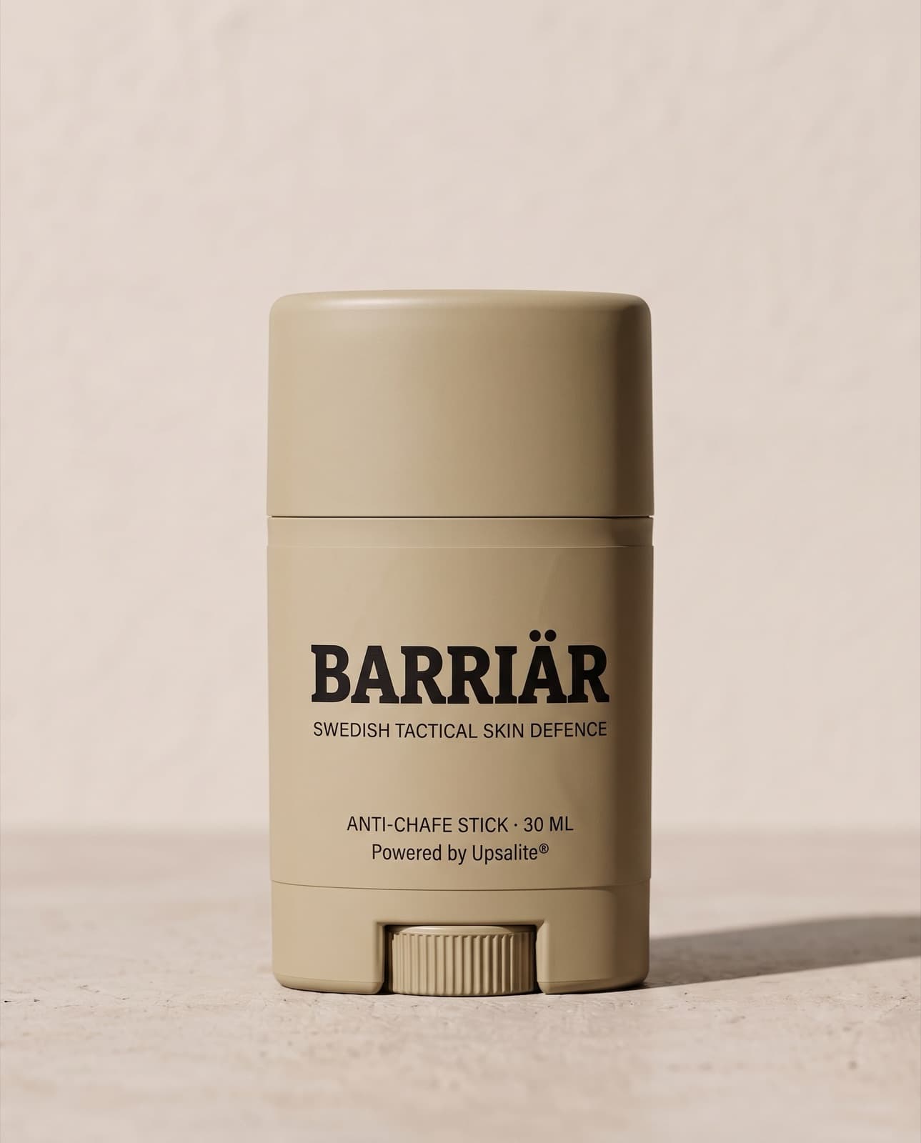 Barriär Anti-Chafe Stick, a short 25 ml sand-beige twist-up stick printed with the Barriär wordmark in Bitter slab-serif over Swedish Tactical Skin Defence, standing on pale concrete against a plaster wall