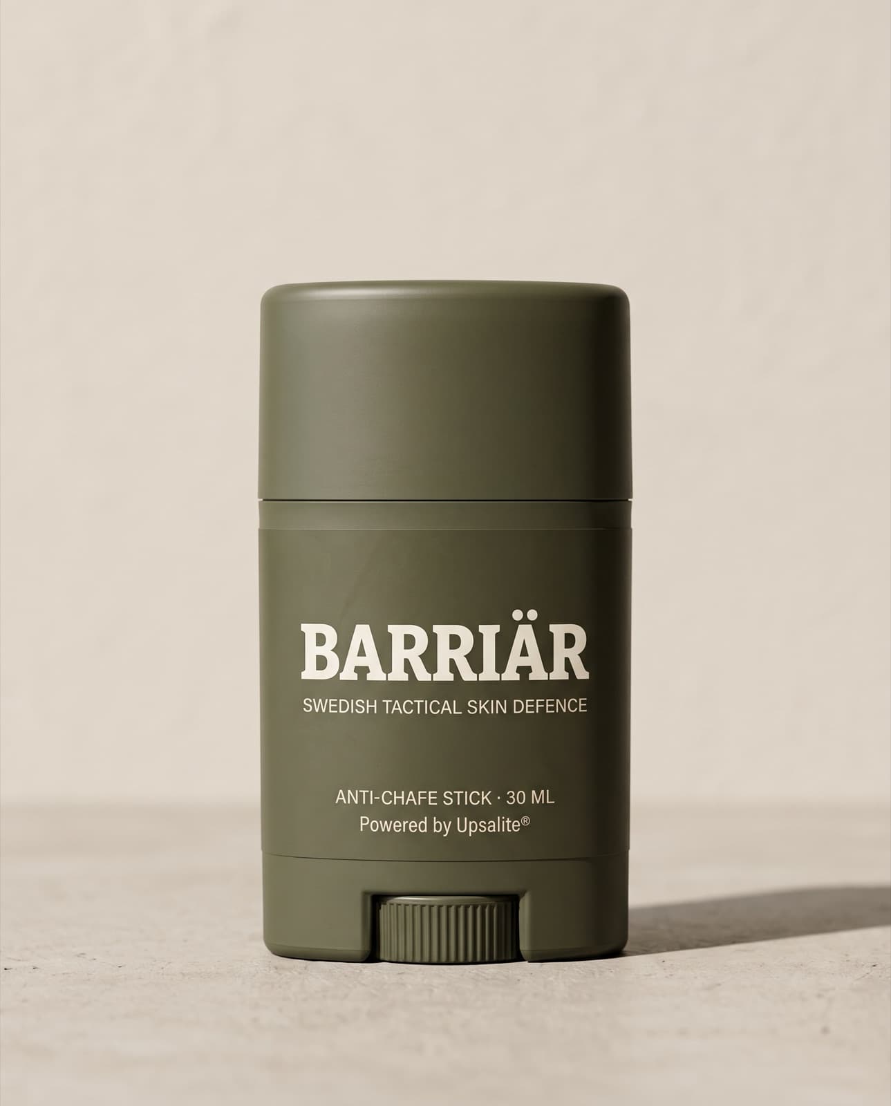 Barriär Anti-Chafe Stick, a short 25 ml olive-green twist-up stick printed with the Barriär wordmark in Bitter slab-serif over Swedish Tactical Skin Defence, standing on pale concrete against a plaster wall
