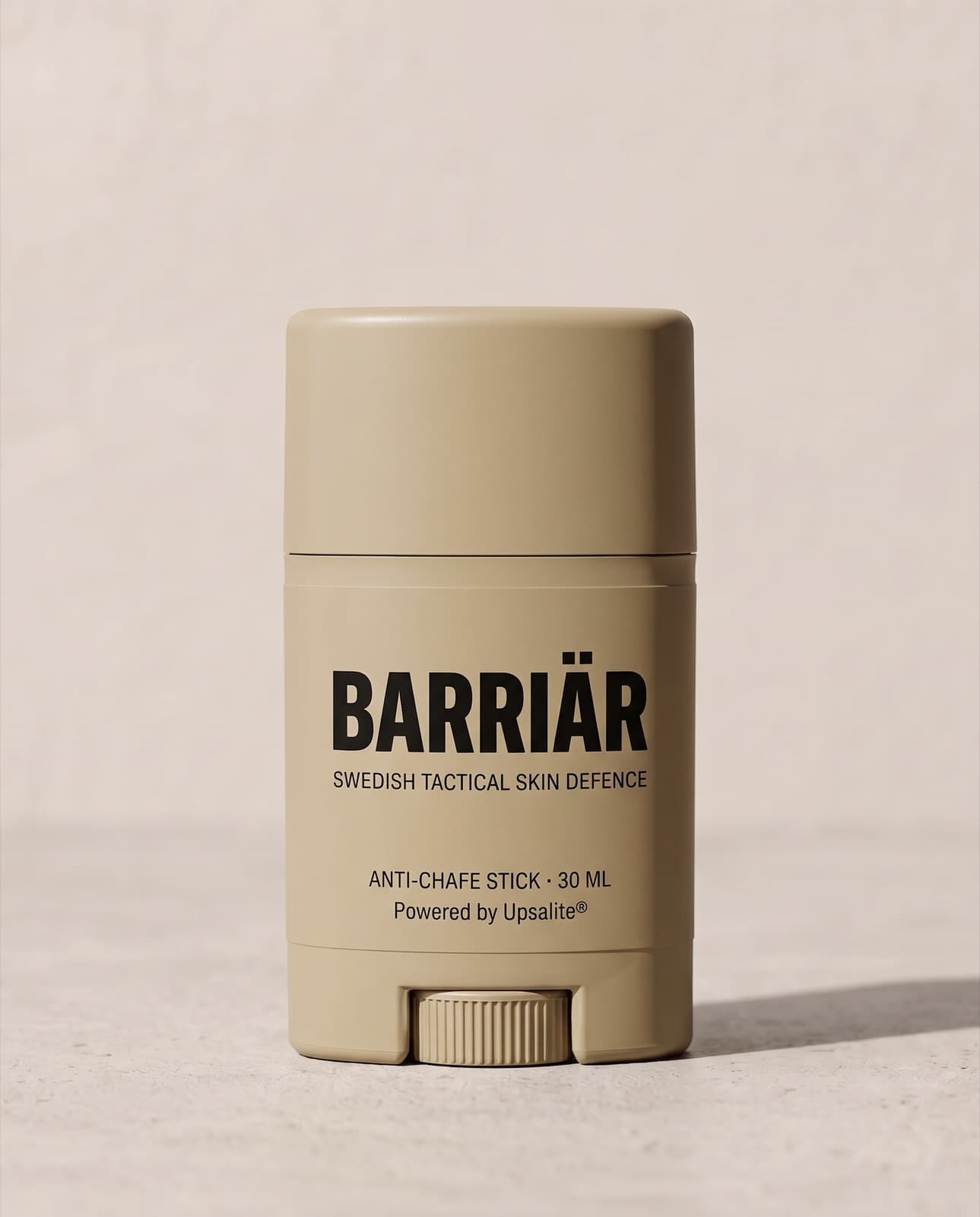 Barriär Anti-Chafe Stick, a short 25 ml sand-beige twist-up stick printed with the Barriär wordmark in Saira condensed over Swedish Tactical Skin Defence, standing on pale concrete against a plaster wall