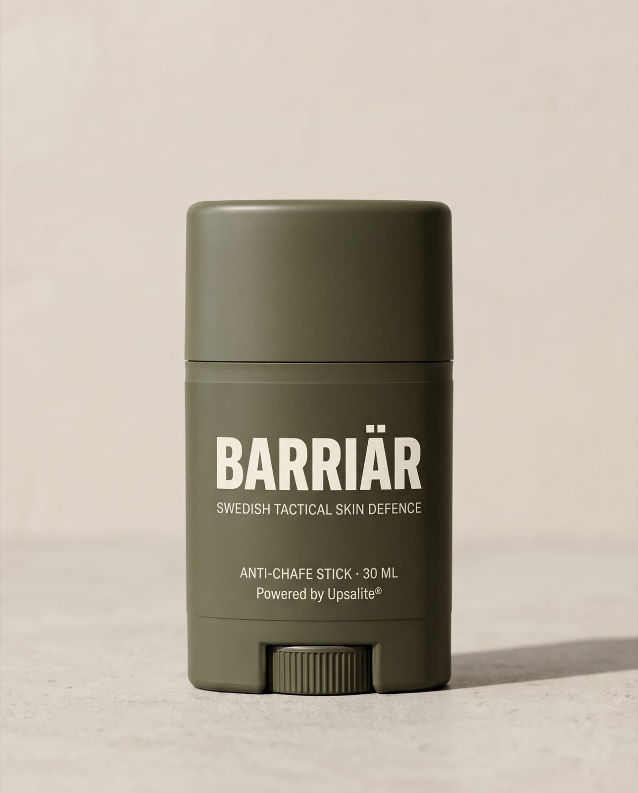 Barriär Anti-Chafe Stick, a short 25 ml olive-green twist-up stick printed with the Barriär wordmark in Saira condensed over Swedish Tactical Skin Defence, standing on pale concrete against a plaster wall