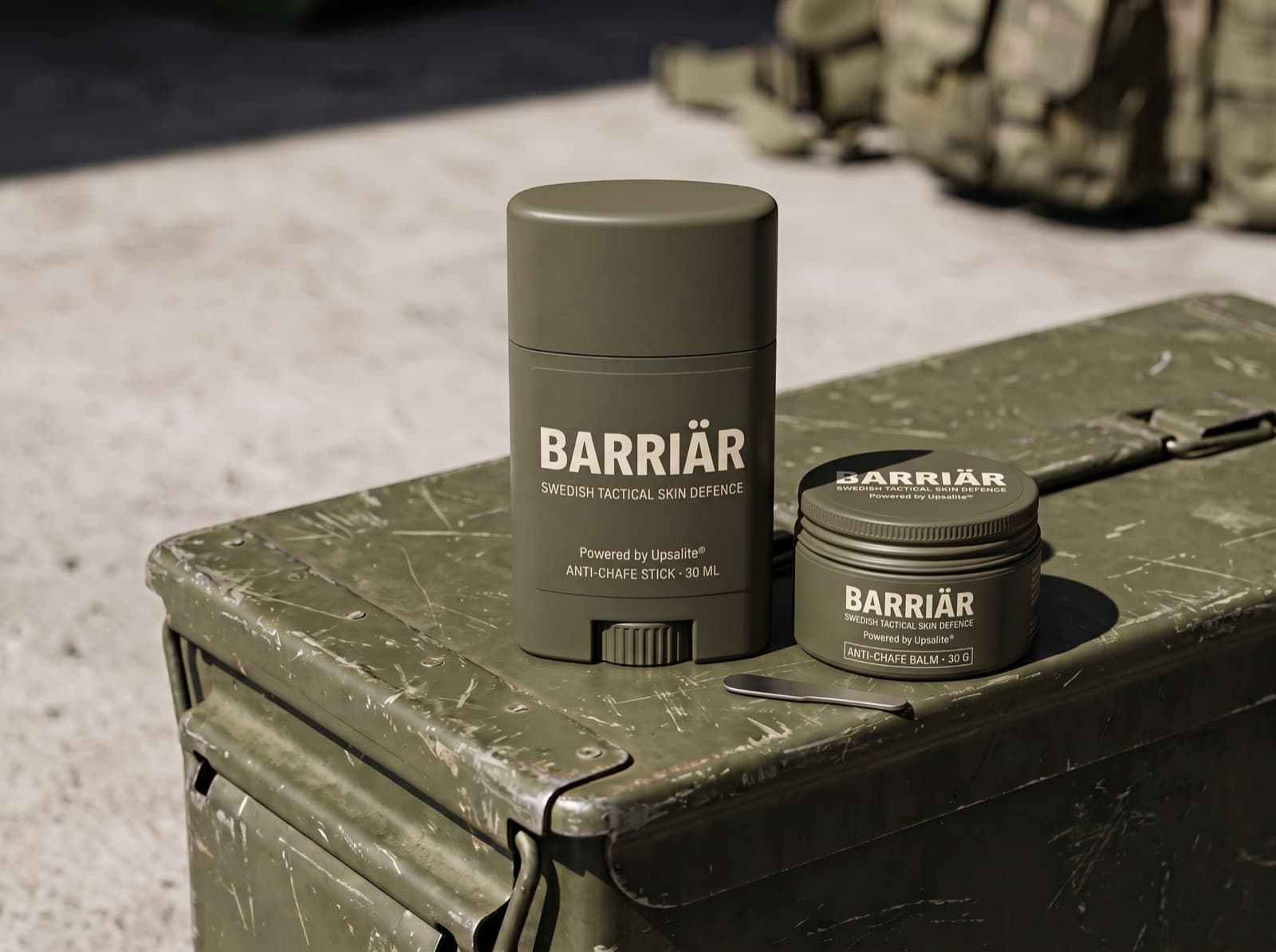 Barriär anti-chafe stick and jar in matte olive packaging on a scratched olive-green ammunition box in hard sunlight, both printed with the Barriär wordmark in Barlow condensed over Swedish Tactical Skin Defence